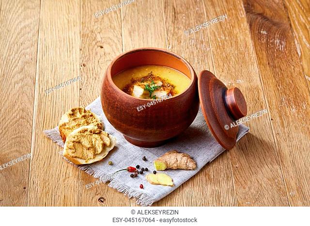 Clay pot of spicy creamy pumpkin soup garnished with dill, served with toasted baguette bread slices covered with paste, ginger on rustic background, close-up
