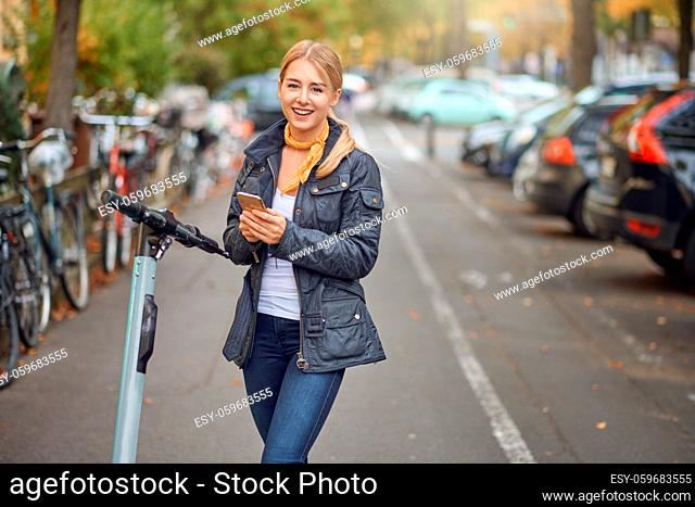 Happy young woman in casual grey jacket looking at camera and smiling, standing in the street with an e-scooter with mobile phone in her hands