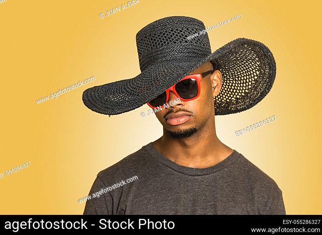 Young African american guy in feminine hat and stylish sunglasses against yellow background