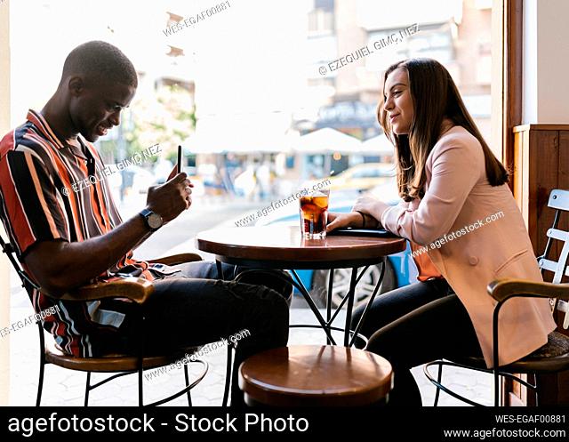 Boyfriend photographing girlfriend through smart phone while sitting at table in cafe