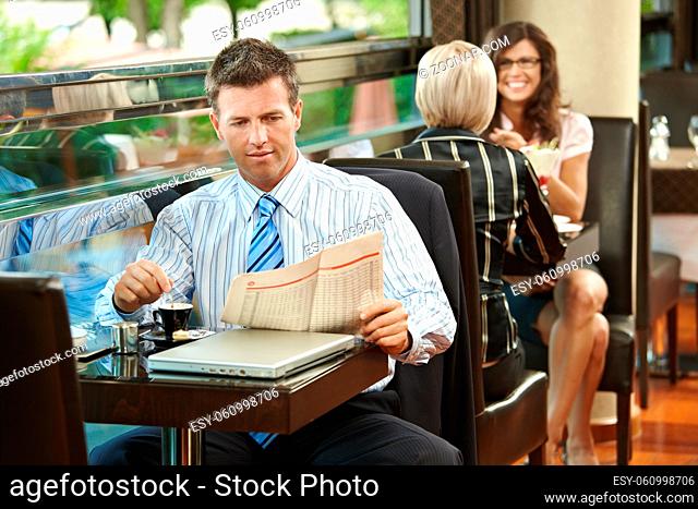 Businessman sitting at table in cafe, reading newspaper and drinking coffee. Young women talking in the background