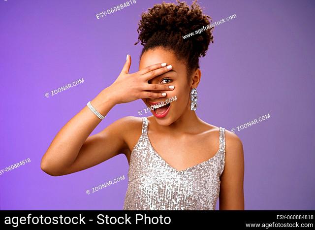 Close-up stylish african american woman afro hairstyle brilliant accessorise glittering silver dress hiding face peeking through fingers amused look surprised...