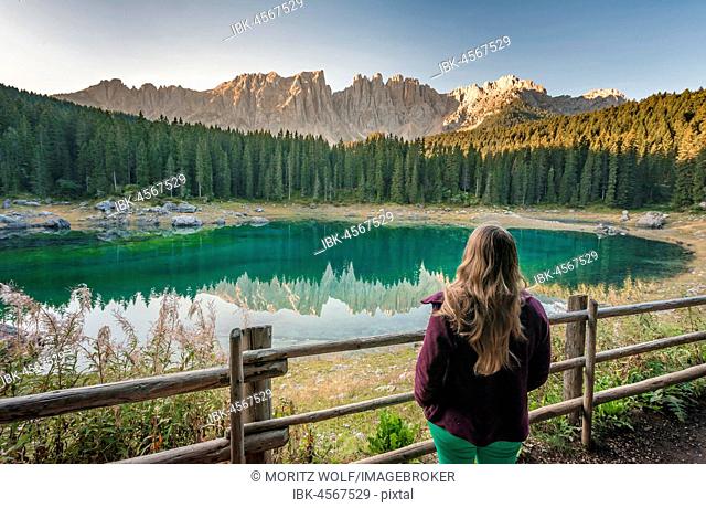Woman overlooks Lake Carezza, Lago di Carezza, morning sun, Latemar group reflected in the lake, mountain tops Torre Diamantidi, Eastern Latemar peak, Col Coron