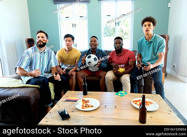 Curious male multiracial friends with beer bottles and pizza on table watching soccer match at home