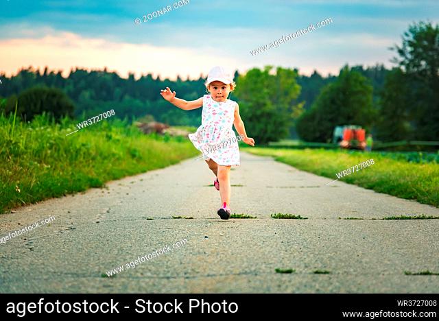 Baby girl with cap outdoors running on a rural road leading from forest