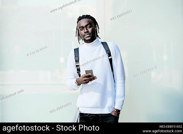 Creative businessman with hands in pockets holding mobile phone against white wall