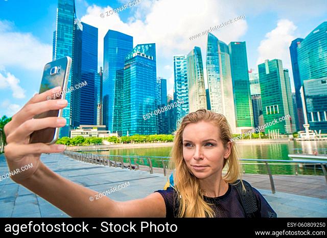 Girl holding smartphone for self-portrait photo with view of modern skyscrapers during summer travel vacation