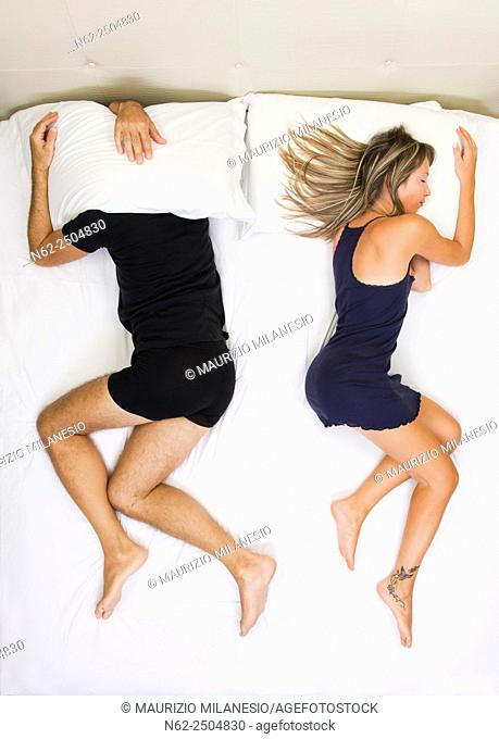 Unhappy couple sleeping far away from each other on bed, him has his head under the pillow, top view