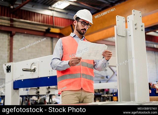 Engineer in protective workwear reading document standing by machinery at factory