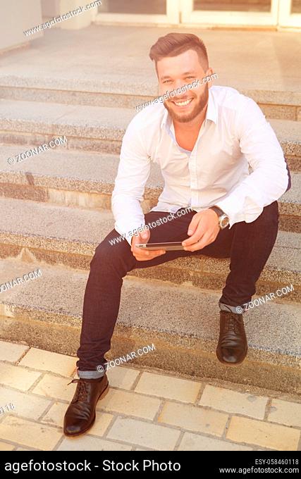 Young smiling man with tablet pc sitting on concrete stairs. Hipster business man in white shirt and black jeans working outdoors. Toned image