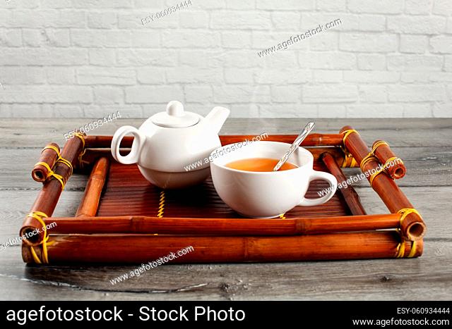 Small white ceramic teapot and cup of hot steaming black tea with silver spoon served on old bamboo tray. placed on gray wood desk