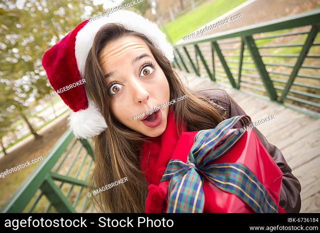 Pretty festive smiling woman wearing a christmas santa hat with wrapped gift and bow outside