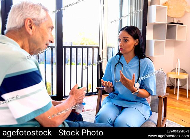 Biracial female physiotherapist talking with caucasian senior man while sitting on chair at home