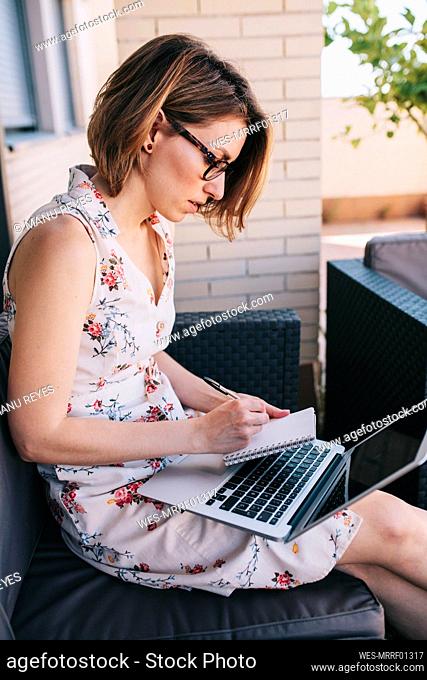 Businesswoman writing in note pad while sitting with laptop on sofa