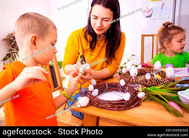 Young mother supporting her sons helping them to create their own Easter decorations at home. Homestyle Easter workshop. Wreath made of vines, Easter eggs