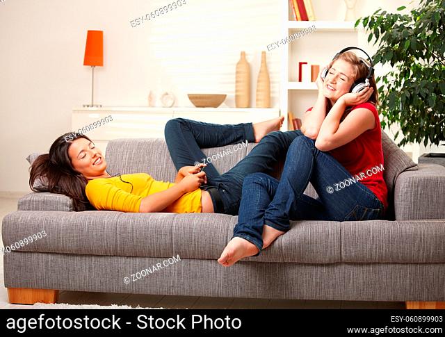 Teen girls listening music on earphones and headphones sitting together on couch at home with closed eyes smiling