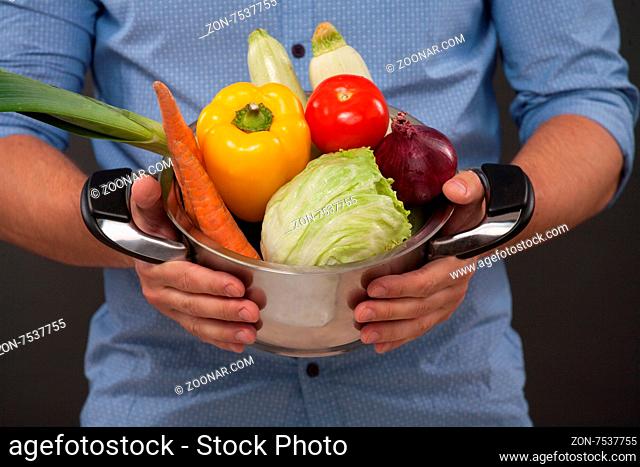 Man keeping pan with fresh vegetables isolated on dark grey. Tomatoe, cabbage, carrot, onion, Bulgarian pepper are ready to be cooked