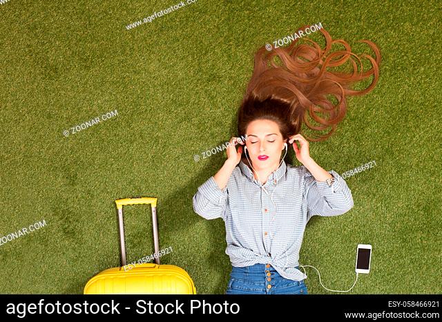Portrit of beautiful tourist lady lying on green grass and listening to music in earphones. Happy woman with long brown hair lying near yellow suitcase