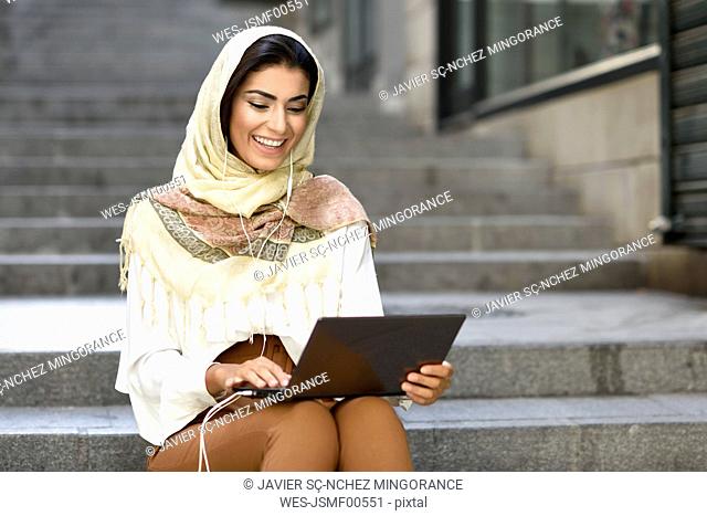 Spain, Granada, young muslim woman wearing hijab using laptop sitting on urban steps