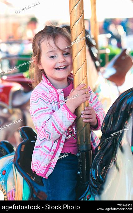 Little adorable smiling girl riding a horse on roundabout carousel at funfair
