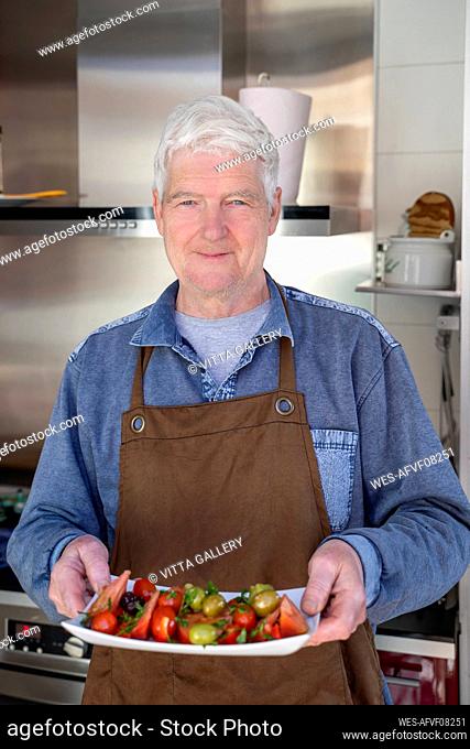 Senior man with apron holding tray of vegetables at kitchen