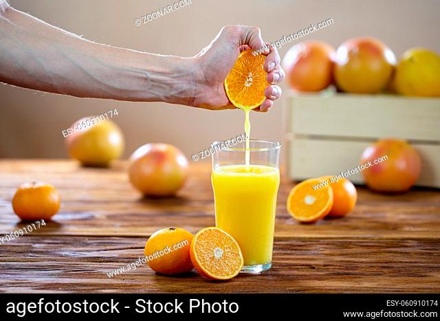 Hand squeezing orange juice into a glass on a wooden table. Human pressing yellow liquid out of fruit. Concept of healthy eating of fresh food