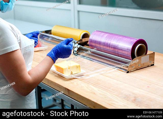 Food packaging. Woman in gloves, apron and mask wraps piece of cheese lying on plastic tray in transparent food film. Concept of food packaging in supermarkets...