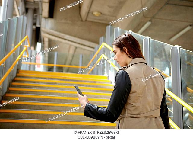 Rear view of young businesswoman reading smartphone texts on stairway, London, UK