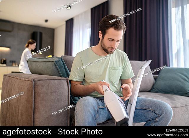 Home appliances. Young adult man looking at detail of vacuum cleaner in hands sitting on sofa and woman behind