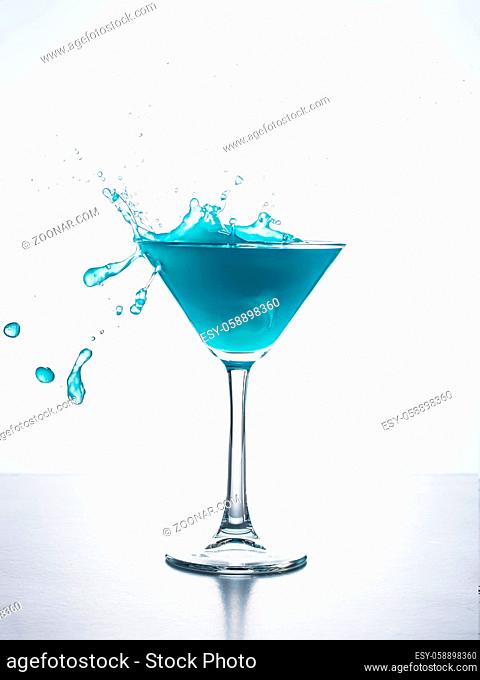 Blue cocktail in martini glass with ice cube splashing into liquid against white background. Blue curacao cocktail with splash on white