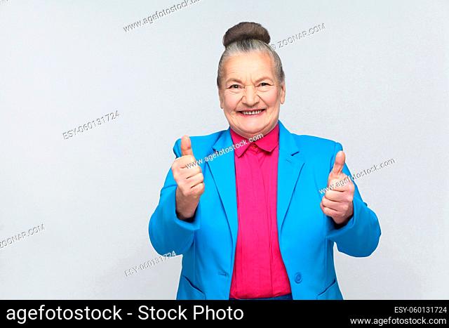 Aged woman toothy smiling with like sign. Emotion and feelings concept. expressive grandmother with light blue suit and pink shirt, collected bun gray hair