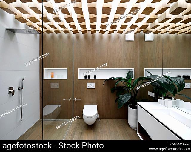 Luminous modern bathroom with design ceiling and wooden walls. There is a glass shower cabin with light tiled wall, white toilet, sink with a mirror