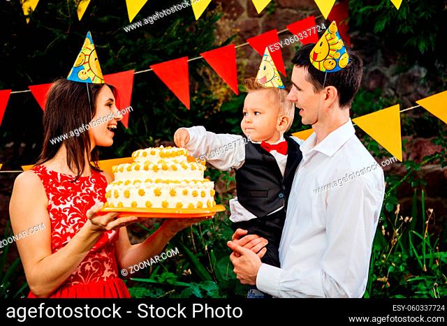 Subject children's birthday party, food and sweets. A young family celebrates one year of son. Dad is holding a big cake, mom is holding a baby in her arms