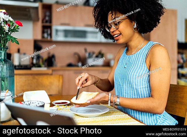 Young woman with curly hair buttering bread on table at home