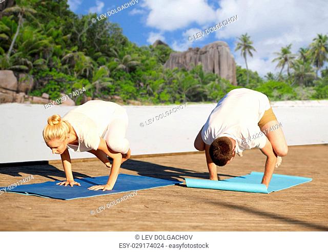 fitness, sport, yoga, people and healthy lifestyle concept - couple making side crow pose on mat over tropical beach background