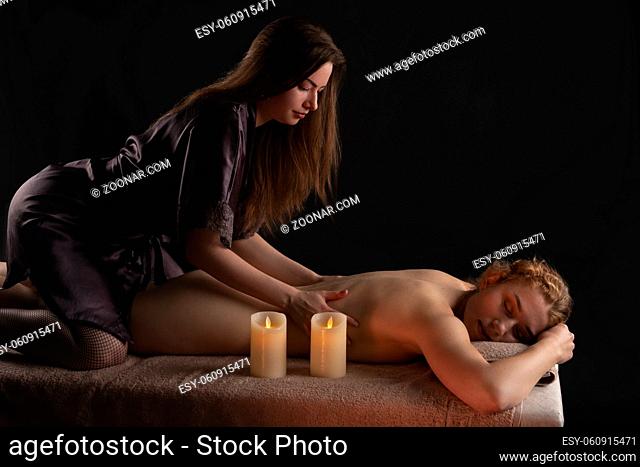 Side view of masseuse massaging shoulders of smiling female client relaxing on table with candle in dark spa salon