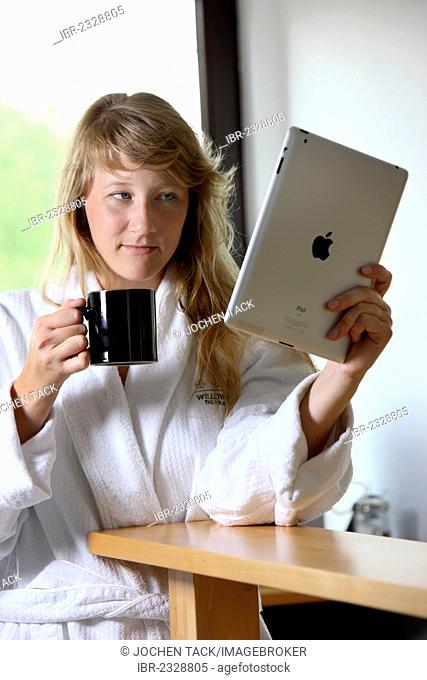 Young woman in the morning in the kitchen, in bathrobe and with a cup of coffee, reading on an iPad, tablet computer, an on-line newspaper