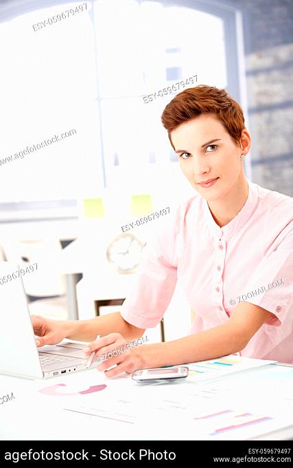 Young businesswoman at work, using laptop computer, holding pen, looking at camera