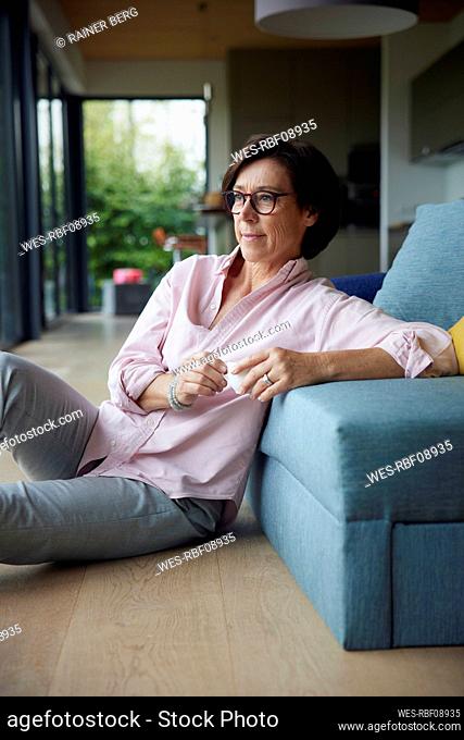 Thoughtful woman with coffee cup sitting on floor by sofa at home