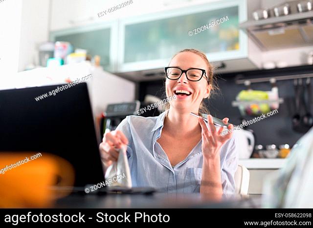 Stay at home and social distancing. Woman in her casual home clothing working remotly from kitchen dining table. Video chatting using social media with friend