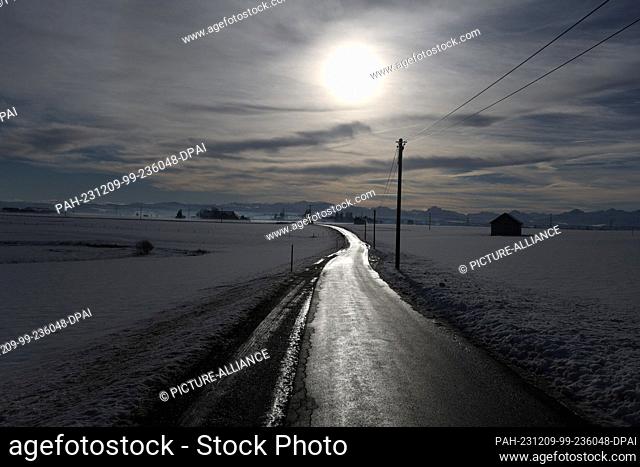 09 December 2023, Bavaria, Ruderatshofen: A road in a wintry landscape glows in the sunshine against the panorama of the Alps