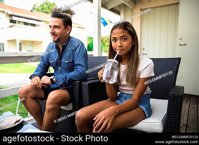 Daughter sipping drink sitting on chair with father at porch