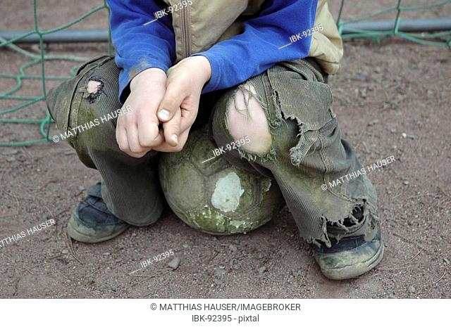 A Boy wearing torn trousers is stitting on a football