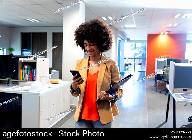 Smiling hispanic businesswoman walking while using smartphone at modern workplace