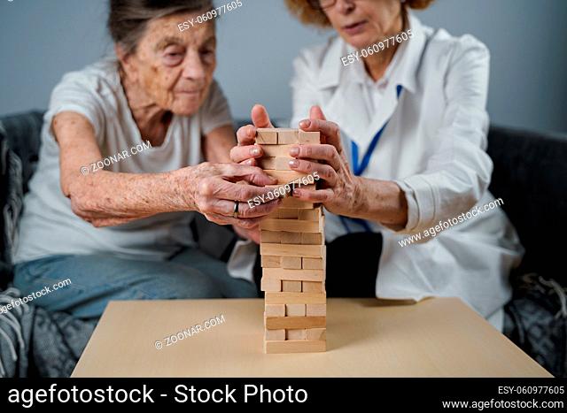 Senior woman playing Jenga, build tower of blocks. Elderly doctor in white lab coat, supporting senior patient, developing logic, fine motor skills