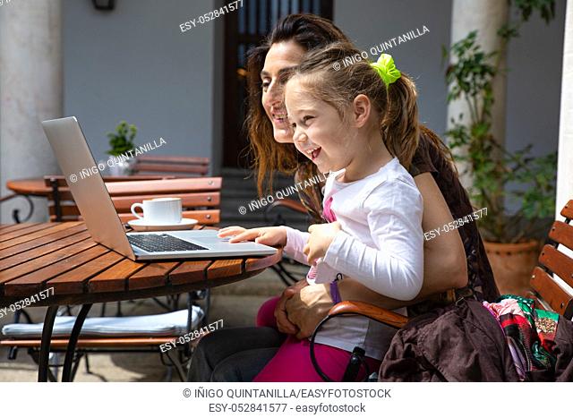 funny girl four years old sitting on mother legs, surfing internet and laughing watching laptop pc computer, with coffee cup on brown wooden table