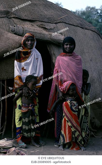 Afar women and children standing outside entrance of domed hut behind