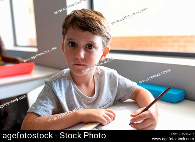 Portrait of caucasian elementary schoolboy with wide eyes sitting at desk in classroom
