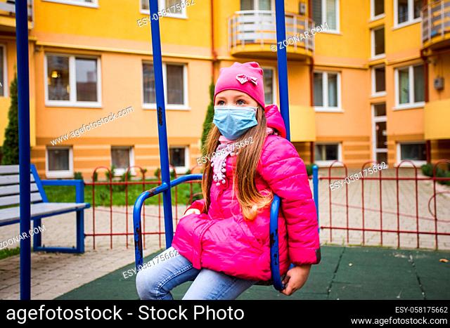 The elementary school girl is wearing a medical mask sitting on a swing in the playground. The girl is wearing a pink jacket and hat in the autumn season