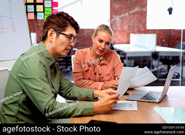 Diverse male and female colleague sitting at table with laptop and paperwork discussing